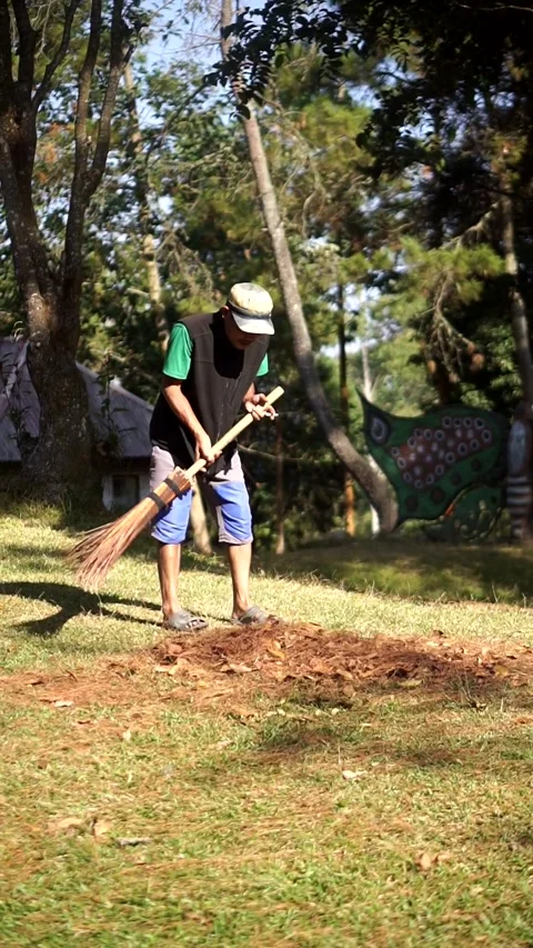 A worker gathers piles of dry leaves using a broom and a wheelbarrow in a park Vídeo Stock 342807063