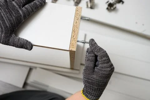 Worker in gloves inserting cam lock screw into white wooden panel close up. Photos