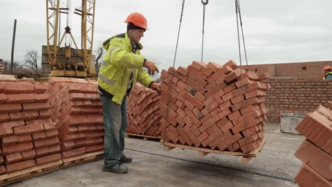Worker guiding a crane lifting a pallet of bricks, A construction worker in Stock Footage 301557699
