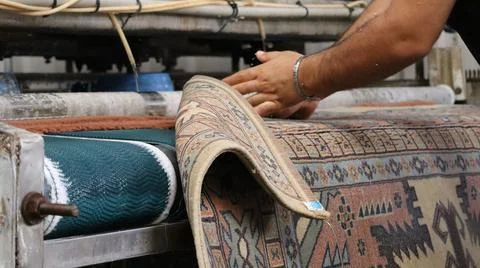 Worker guiding patterned rug through industrial cleaning rollers in workshop Stock Photos