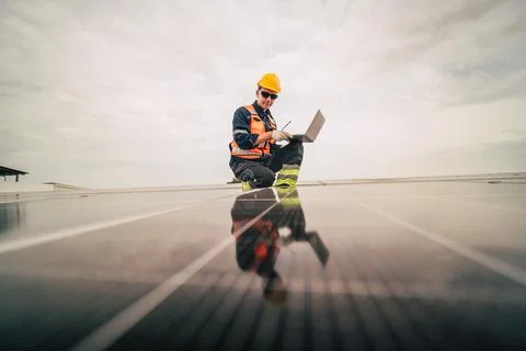 Worker inspects solar panels on rooftop while using a laptop in daylight hour Stock Photos