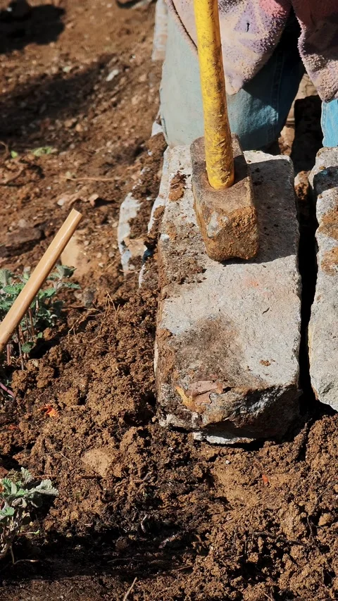 Worker laying heavy stone block in dirt, compacting soil with a hammer for solid Stock Footage 332164407
