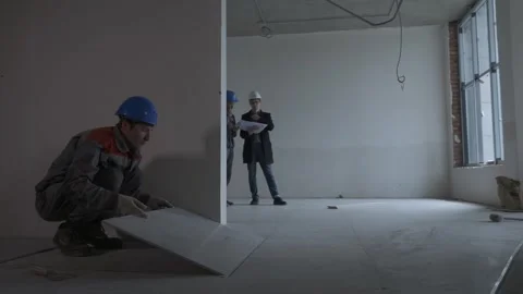 Worker laying tiles. In the next room, engineers are discussing Stock Footage 321004567