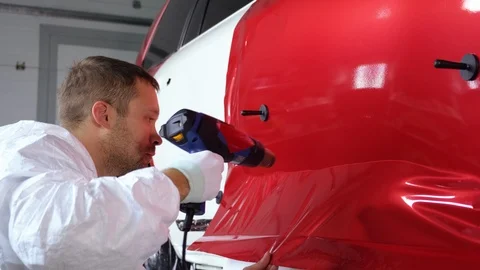 Worker man is warming vinyl tape by fan and fixing on door of automobile Stock Footage 121416038