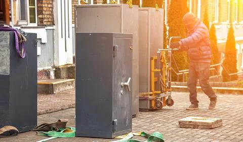 A worker is moving a large safe with a dolly in a city setting during the aft Stock Photos