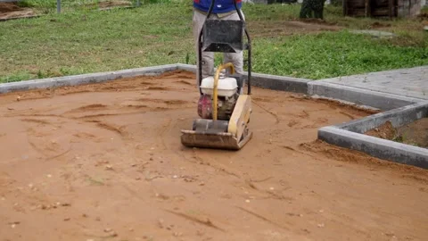 Worker operates plate compactor on leveled muddy ground beside concrete borders Stock Footage 344874114