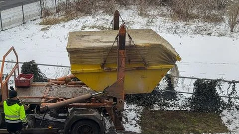 A worker operates a yellow skip loader truck to unload a large metal container Stock Photos