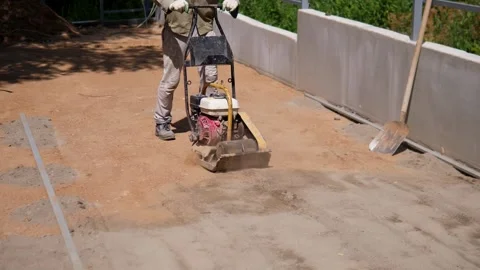 Worker operating plate compactor on dusty construction ground beside retaining Stock Footage 344877726