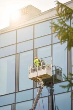 A worker in overalls on an elevator washes the glass wall of a building. Stock Photos