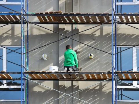 A worker plastering a building facade while standing on a scaffold. Construct Stock Photos