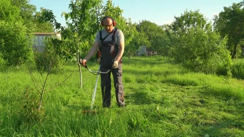 Worker in protective gear trims grass with a string trimmer in a lush garden, 動画素材 337374480