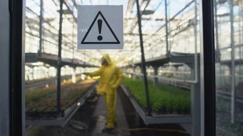 Worker in protective uniform hosing plants in greenhouse, warning sign on door Видео 118934798