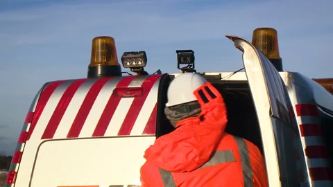 A worker in a red jacket by the car. The car with flashing lights is at the Stock Footage 94487265