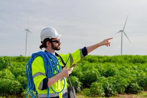 Worker in reflective gear points at wind turbines in a green field while ho.. Stock Photos