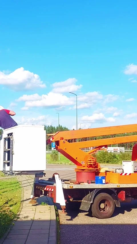 Worker repairs equipment on a platform lift near a busy road during a clear day Stock Footage 325312931