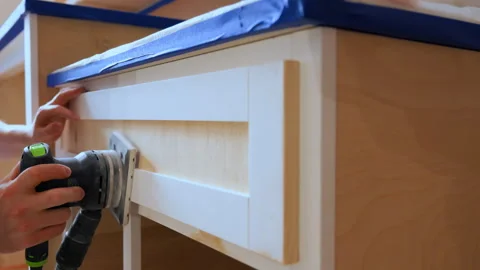 Worker sanding wooden cabinet surface with an electric orbital sander during Stock Footage 334019892