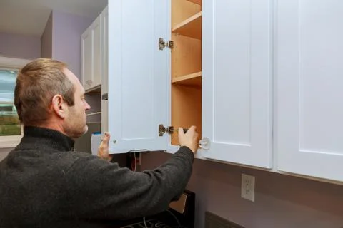 Worker sets a new handle on the white cabinet with a screwdriver installing k Stock Photos