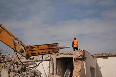 A worker stands on a collapsed building next to an excavator's hydraulic hamm 库存照片