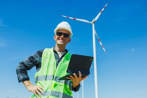Worker stands in front of wind turbine holding laptop while wearing safety .. Stock Photos