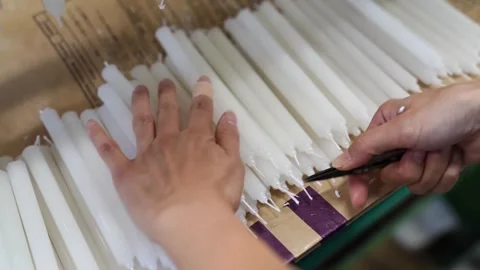 Worker trimming wicks at the base of tapered candles laid on a paper covered Stock Footage 343875734