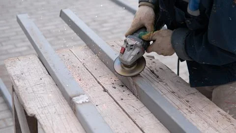 Worker using angle grinder on metal beams beside wooden workbench, creating Stock Photos
