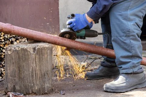 Worker using an electric angle grinder to cut a rusty metal pipe on a log i.. Stock Photos