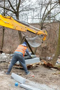 A worker using an excavator bucket unloads pallets of concrete curbs. Stock Photos