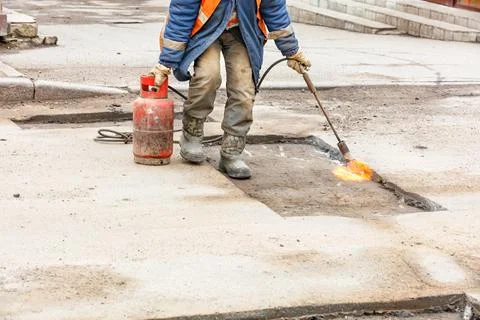 A worker using a gas burner heats the asphalt pavement for patching the road. Stock Photos
