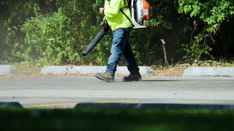 Worker using a leaf blower to clear fallen leaves and debris along edge of a Stock Footage 319724664