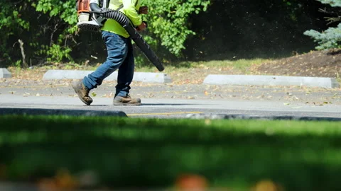 Worker using a leaf blower to clear fallen leaves and debris along edge of Stock Footage 320264429