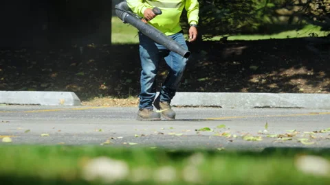 Worker using a leaf blower to clear fallen leaves and debris along the edge of Stock Footage 320448362