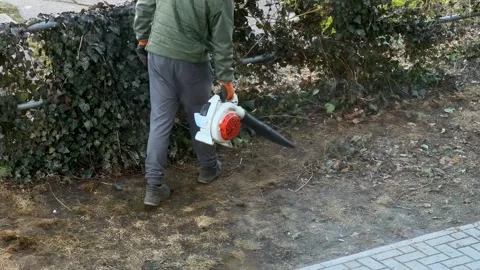 A worker using a leaf blower to clear debris near a green garden fence, Stock Footage 346483371