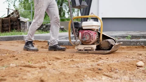 Worker walking beside plate compactor on sandy construction site with concrete Stock Footage 344873786