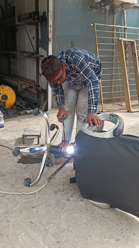 A worker welding a broken office chair in a workshop, wearing safety glasses whi Stock Footage 308621928