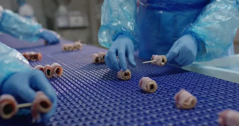 Workers in blue gloves place stuffed bacon rolls onto skewers at a blue 動画素材 339990134