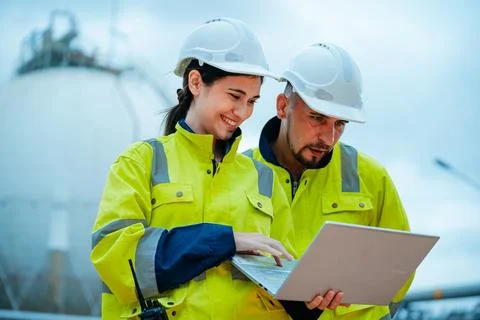 Workers collaborate on a project while using a laptop at an industrial site.. Stock Photos