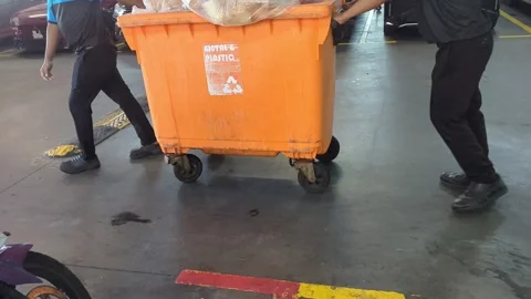 Workers Pushing Large Orange Waste Bin in Indoor Parking Lot Stock Footage 328969482