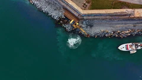 Workers use a digger to remove rocks from the shoreline while a boat floats Stock Footage 331062613