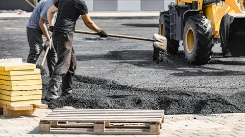 Workers use shovels and a machine to spread asphalt on a ground area. Pallets Stock Photos