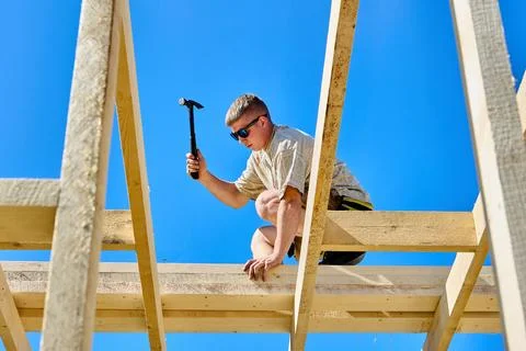 Working at height as young construction worker uses hammer to fasten timber.. Stock Photos