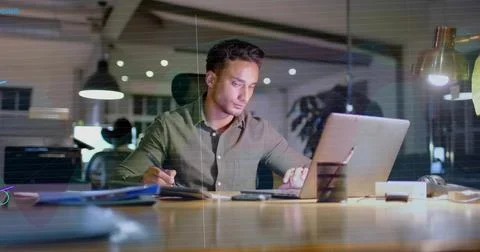 Working Hispanic man wearing olive shirt typing on silver laptop taking notes in Stock Photos