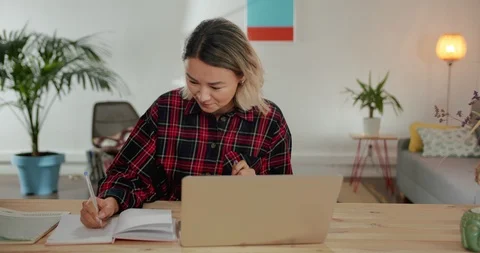 Working process of nice pensive Asian girl taking notes at modern cozy living Stock Footage 120948352