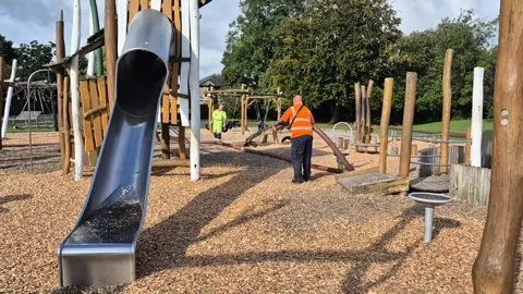 Workman replacing soft woodchip on a children's playground Stock Footage 284555988