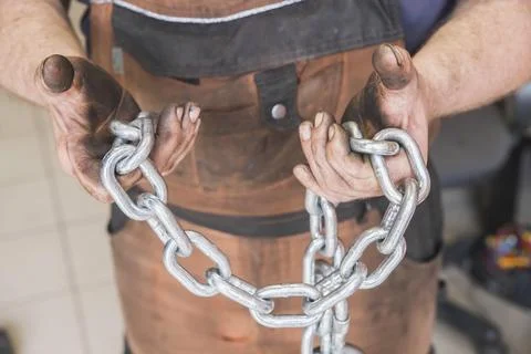 Workshop worker in dirty brown clothes holds steel chain in hands Stock Photos