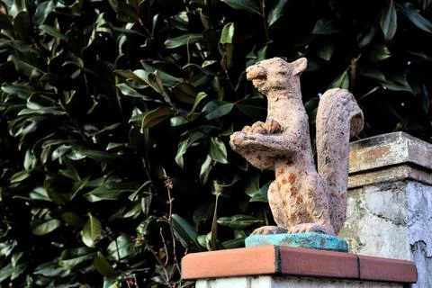 Worn out statue of a squirrel on the top of a gate with a hedge as background Stock Photos
