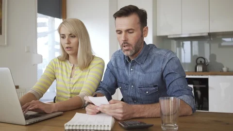 Worried couple sitting at dining table with laptop and bills talking about ho 動画素材 114605842