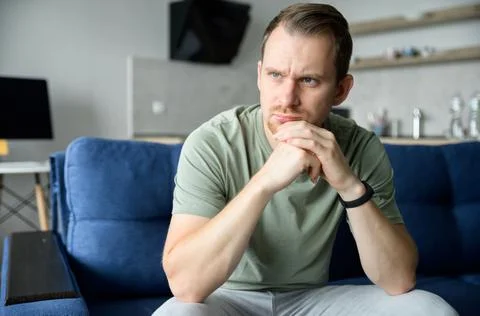 Worried guy sits on the sofa alone and looks away lost in thoughts Stock Photos