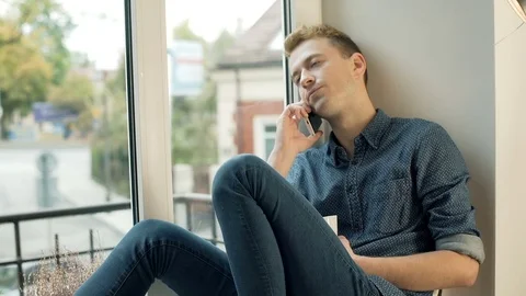 Worried man sitting by the window and talking on cellphone Stock-Footage 70380115