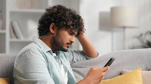 Worried young Indian boy sitting on sofa looking at her phone and does not Stock Photos