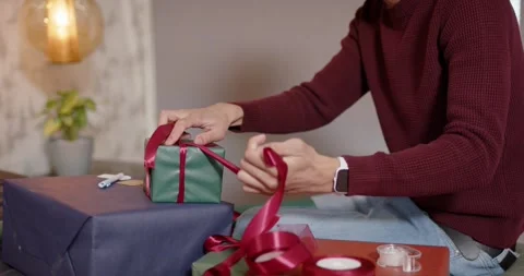 Wrapping at home man in Santa hat unrolling ribbon tying bow on green gift, copy Stock Footage 343468436